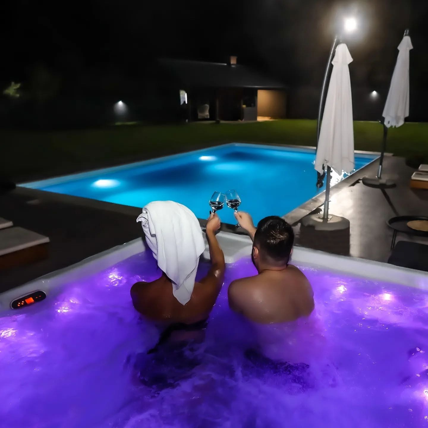 Par u jacuzziju s čašama vina, bazen u pozadini